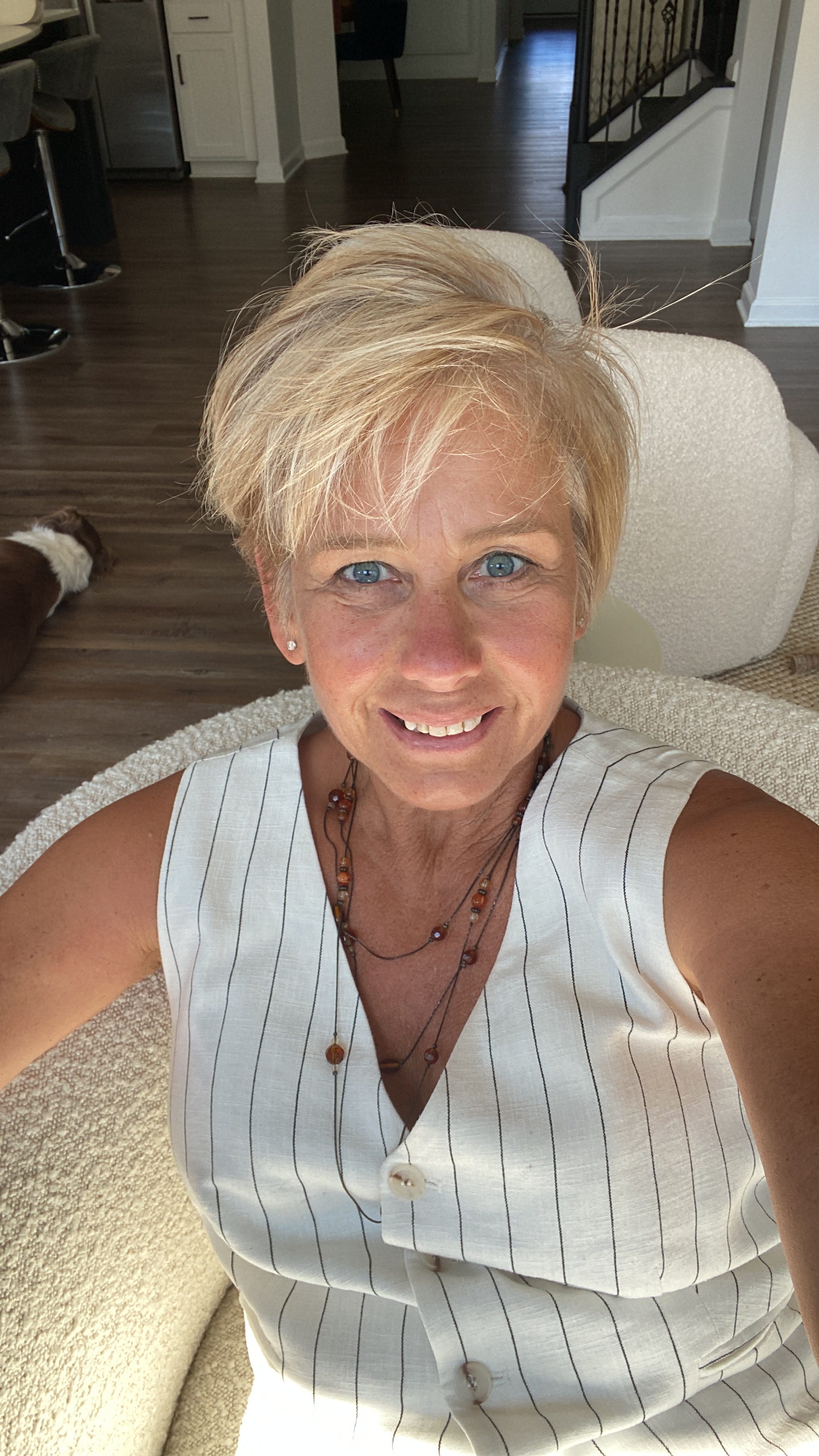 A woman with short blonde hair, wearing a white sleeveless top with black stripes, is smiling at the camera.